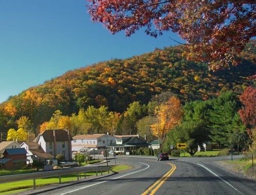 A scenic road curves through a small town with colorful autumn foliage on the surrounding hills.