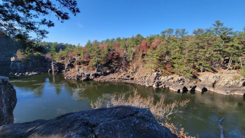 A serene river flows through rocky terrain, surrounded by trees with autumn foliage under a clear blue sky.