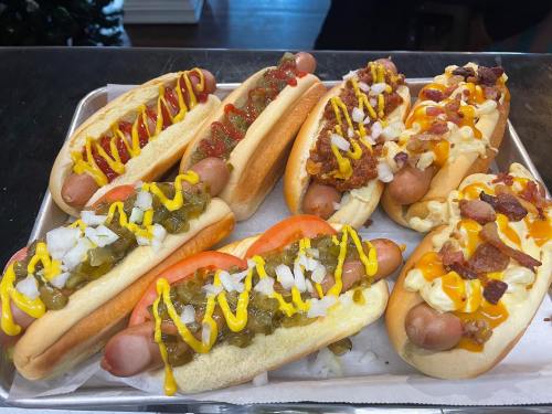 A tray of eight gourmet hot dogs with various toppings, including ketchup, mustard, onions, and bacon.