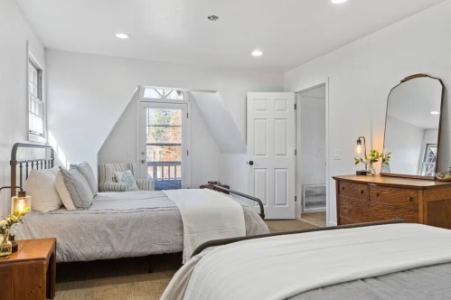 Bright, airy bedroom with two beds, a dresser, and a door leading to a balcony, featuring white walls and natural light.