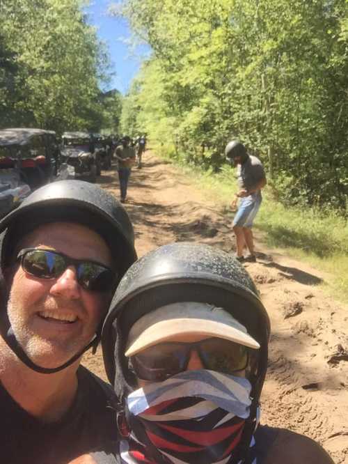 Two people in helmets and masks smile for a selfie on a dirt trail, with others and vehicles visible in the background.