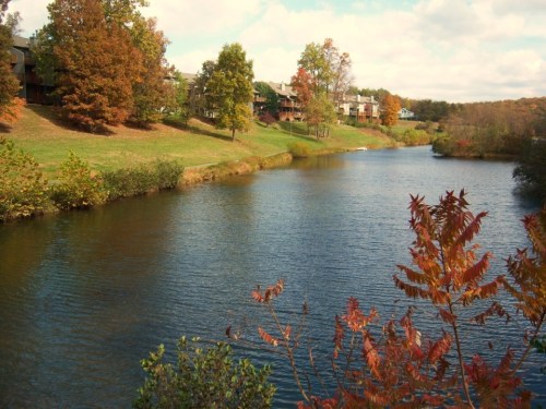 A serene river scene surrounded by autumn trees and buildings in the background under a partly cloudy sky.