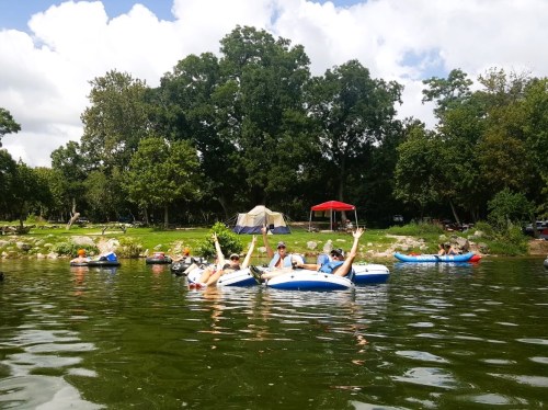 Group of people in inflatable kayaks on a river, enjoying a sunny day with trees and a campsite in the background.