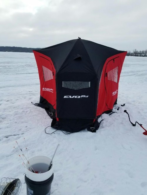 A black and red ice fishing shelter set up on a snowy lake, with fishing gear nearby.