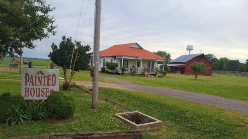 A rural scene featuring a house with a red roof, a barn, and a sign reading "Painted House" amidst green grass.