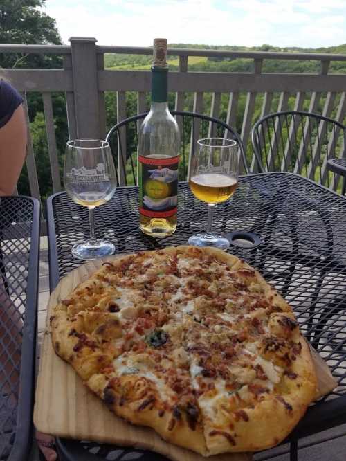 A wooden table with a pizza, two glasses of wine, and a bottle, set against a scenic outdoor backdrop.