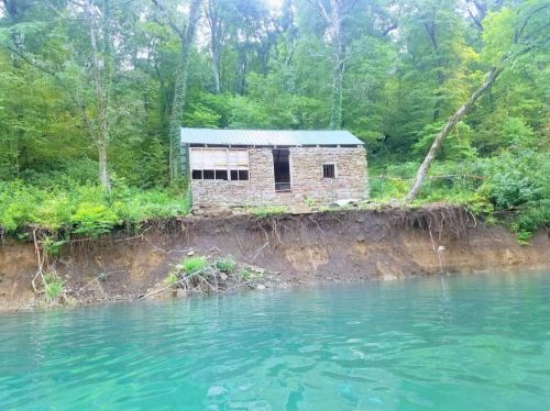 A stone building with a metal roof sits on a riverbank, surrounded by lush green trees and clear blue water.