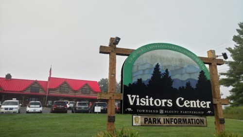 Sign for a Visitors Center with a rustic design, surrounded by greenery and a foggy atmosphere. Cars parked nearby.
