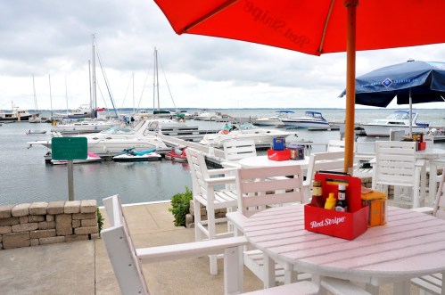 A marina view with boats docked, featuring outdoor seating and umbrellas at a waterfront restaurant.