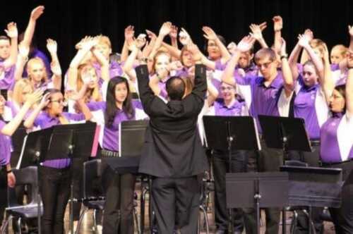 A choir in purple shirts performs on stage, with a conductor leading them and hands raised in unison.