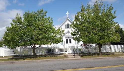 A white church with a cross, surrounded by green trees and a white picket fence, under a blue sky.