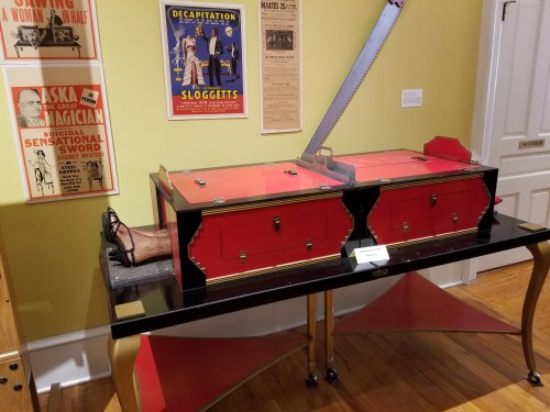 A vintage magician's illusion box with two red compartments, displayed on a black table, alongside vintage posters.