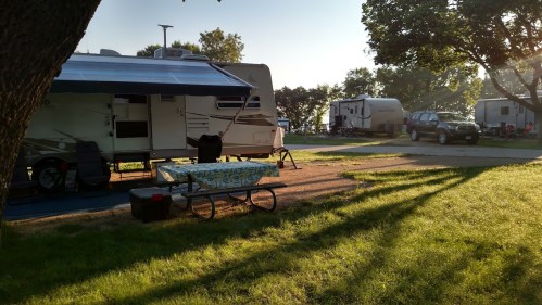 A campsite with an RV, picnic table, and trees, bathed in warm sunlight, with another RV in the background.