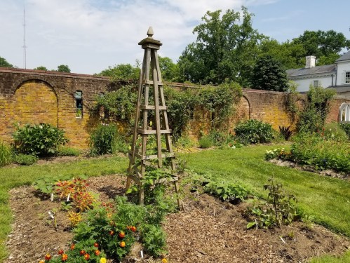 A wooden trellis stands in a lush garden surrounded by brick walls and vibrant flowers under a clear sky.