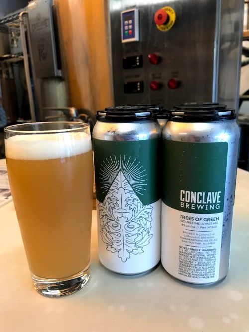 A glass of hazy beer next to two cans of "Trees of Green" from Conclave Brewing on a table.