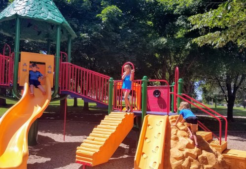 Children playing on a colorful playground with slides, climbing structures, and a rock wall under sunny skies.