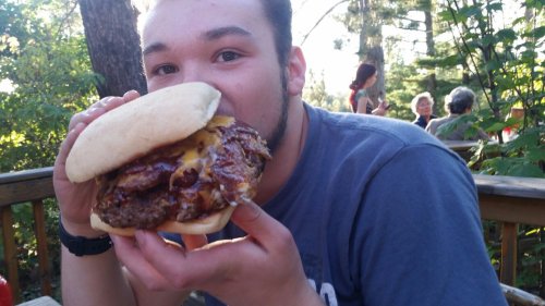 A person holds a large burger with cheese, smiling outdoors in a wooded setting.