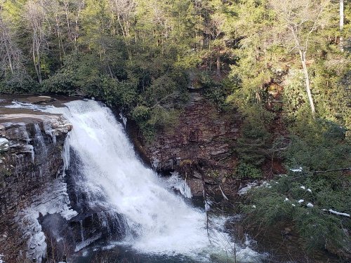A cascading waterfall surrounded by lush green trees and rocky terrain, with some ice visible near the water's edge.