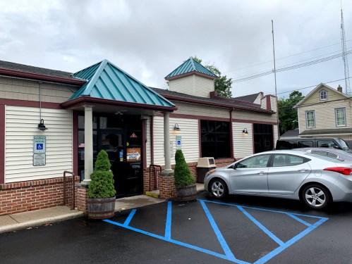 A one-story building with a teal roof, brick accents, and a parking area featuring a gray car and green shrubs.