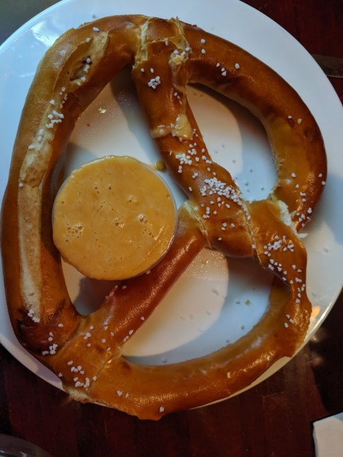 A large, soft pretzel with coarse salt served on a plate alongside a small bowl of cheese dip.