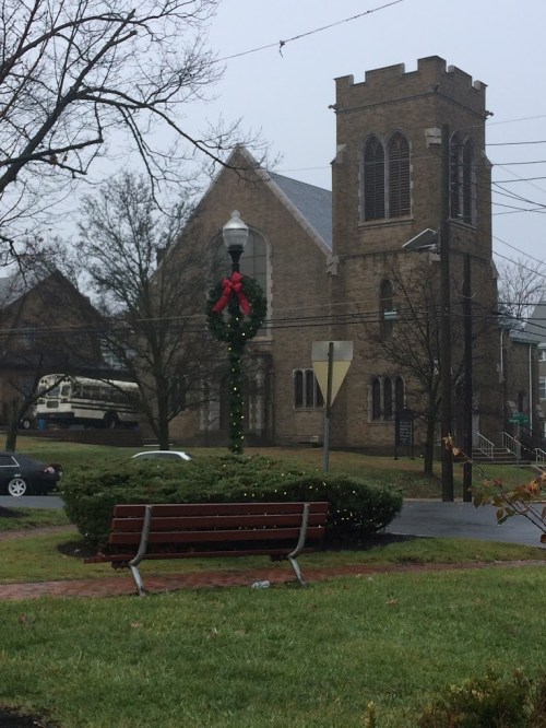 A foggy scene featuring a church with a wreath on its tower, a bench in the foreground, and a bus in the background.