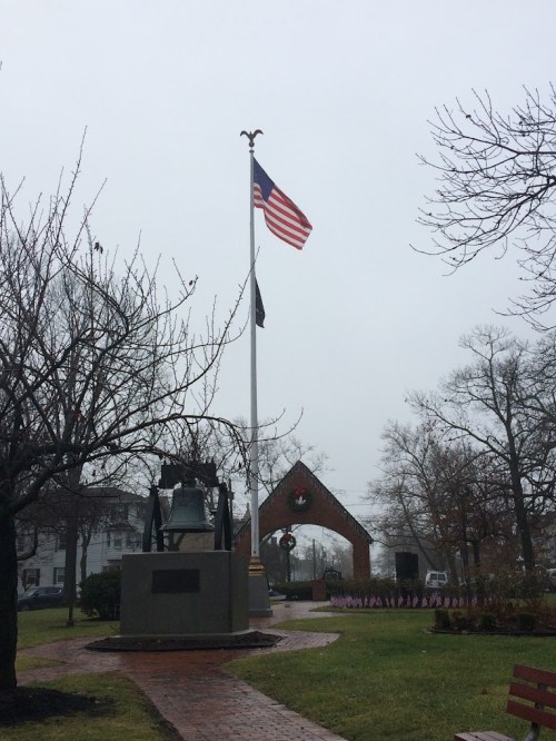 A foggy park scene featuring an American flag, a bell monument, and a decorative archway with holiday wreaths.