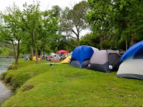 A campsite by a river with various colorful tents set up among green grass and trees.