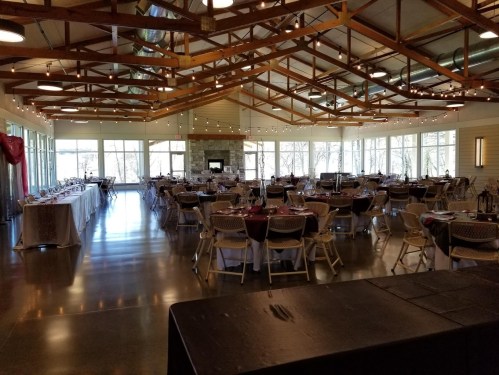 A spacious event venue with tables set for dining, large windows, and a wooden ceiling with exposed beams.