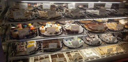 A display case filled with various desserts, including cakes, cupcakes, and brownies, all labeled with names.