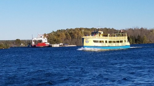 A yellow tour boat navigates blue waters near a red cargo ship and tree-lined shore under a clear sky.