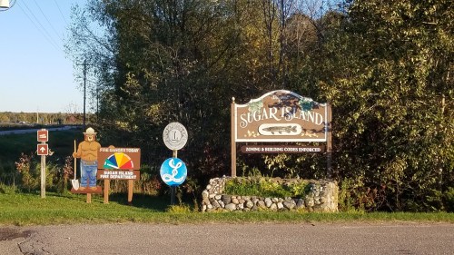 Sign for Sugar Island with a wooden figure, fire department notice, and local organization emblem. Lush greenery in background.