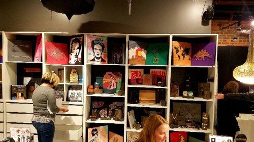 A display of colorful artwork and crafts on shelves, with two people browsing and organizing items in a cozy setting.