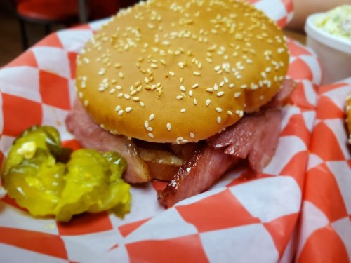A sesame seed bun sandwich with layers of meat, served with pickles on a red and white checkered paper.