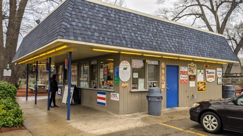 A small, colorful food stand with a blue door, outdoor seating, and various signs, set in a park-like area.