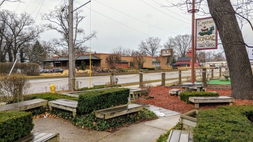 Outdoor seating area with wooden benches and shrubs, near a road with a sign for "Captain Sundae" in the background.