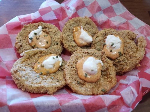A basket of six fried green tomatoes topped with creamy sauce and garnished with herbs on a red and white checkered paper.