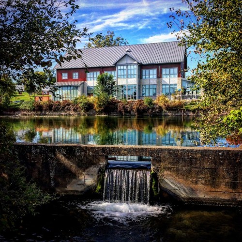 A modern building reflects in a calm river, with a small waterfall in the foreground and greenery surrounding the scene.