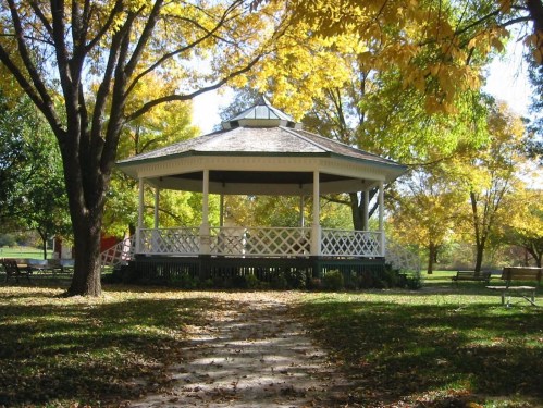A white gazebo surrounded by colorful autumn trees and fallen leaves in a park setting.