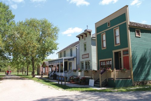 Historic buildings line a dirt path, surrounded by trees and blue skies, with colorful flowers adorning the porches.