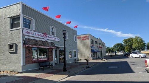 A street view featuring the Great Lakes Popcorn Co. with red flags, alongside other buildings and a clear blue sky.