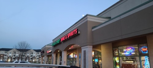 Exterior of a liquor store with neon signs, under a clear sky with a visible moon. Nearby buildings in the background.
