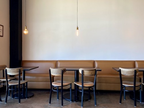 A minimalist café interior featuring wooden tables, chairs, and a brown leather bench against a plain wall.