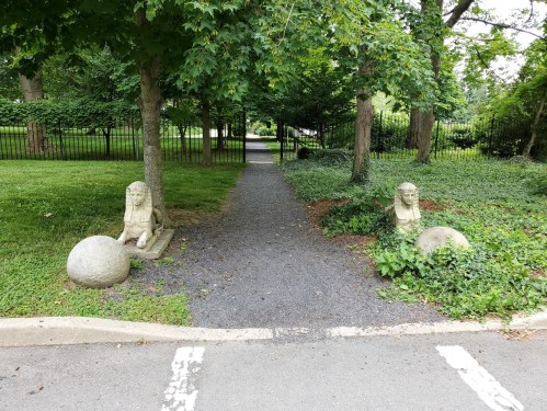 A gravel path lined with trees, featuring two stone sphinx statues and large round stones on either side.