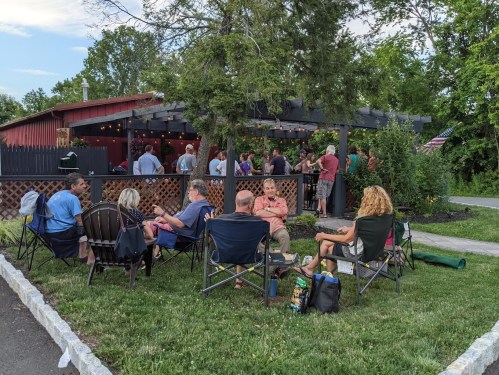A group of people socializing outdoors near a red building, seated in chairs with greenery and string lights around.