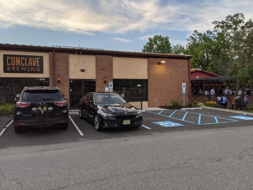 Exterior of Conclave Brewing with parked cars and outdoor seating area, under a clear sky.