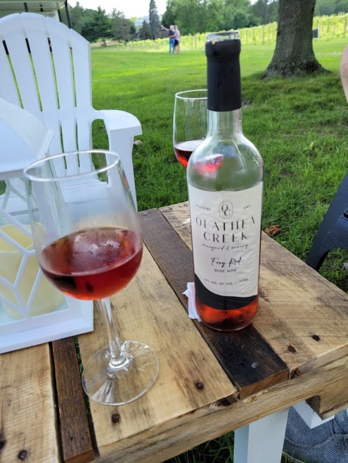 A bottle of Olathea Creek rosé wine with two glasses on a wooden table, set in a vineyard.