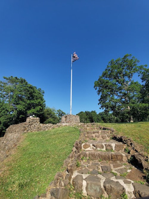 Stone steps lead up to a flagpole on a grassy hill, surrounded by trees under a clear blue sky.