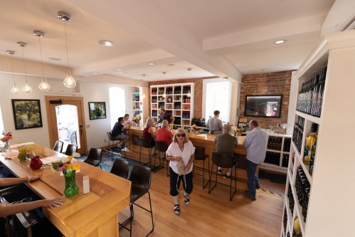 A cozy wine bar with patrons enjoying drinks, a wooden counter, and shelves filled with bottles. Bright and inviting atmosphere.