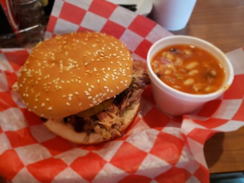 A sesame seed bun sandwich filled with pulled pork, pickles, and a side of hearty bean soup in a white cup.