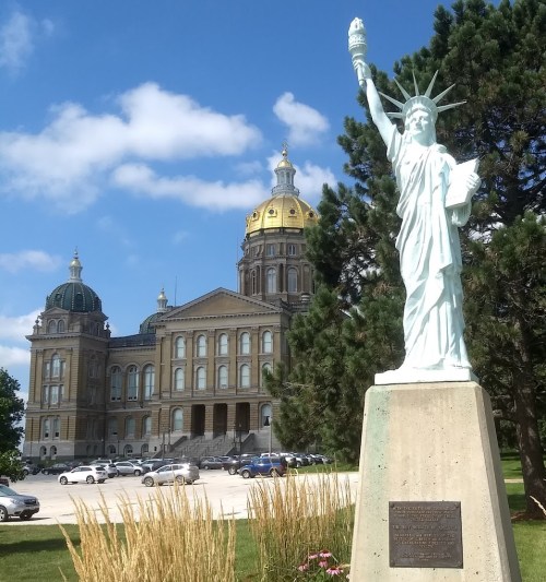Statue of Liberty replica in front of a historic building with a golden dome, surrounded by greenery and blue sky.